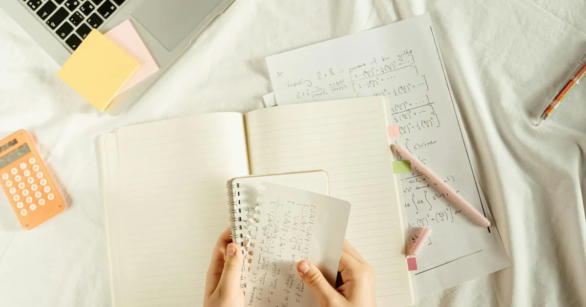 Hands holding a notebook with math equations, calculator, and laptop on a study desk.