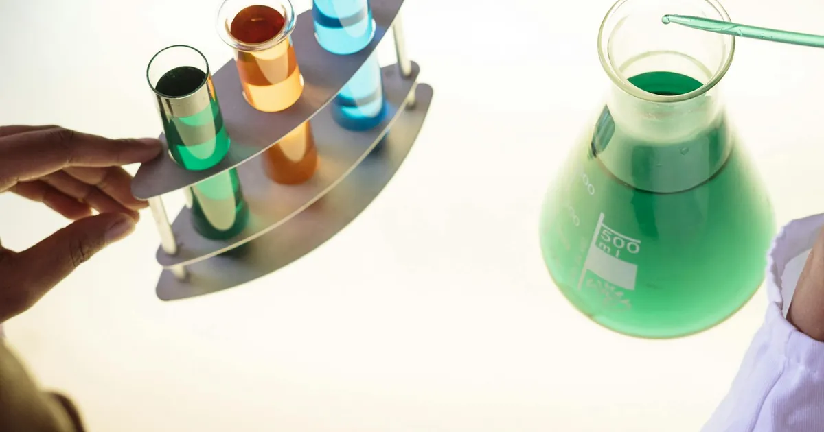 Unrecognizable ethnic scientist in white coat putting tube into flask with liquid substance while conducting chemical experiment with reagents in test tubes in laboratory