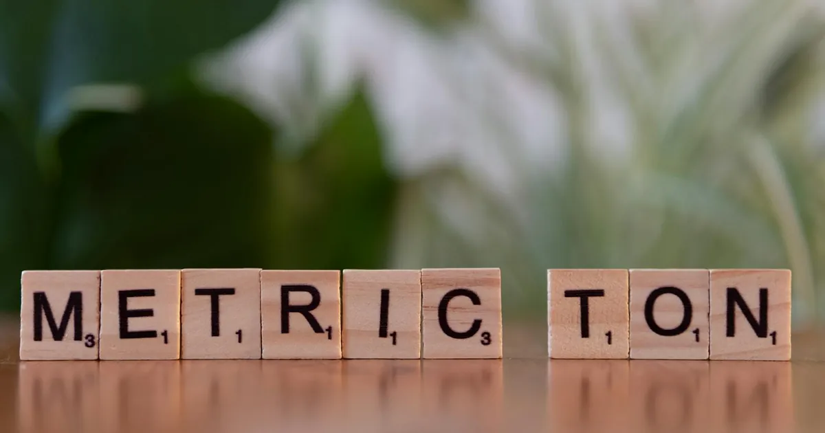 Scrabble tiles on a brown surface spell 'Metric Ton' with blurred green background. Scrabble tiles on a brown surface spell 'Metric Ton' with blurred green background.