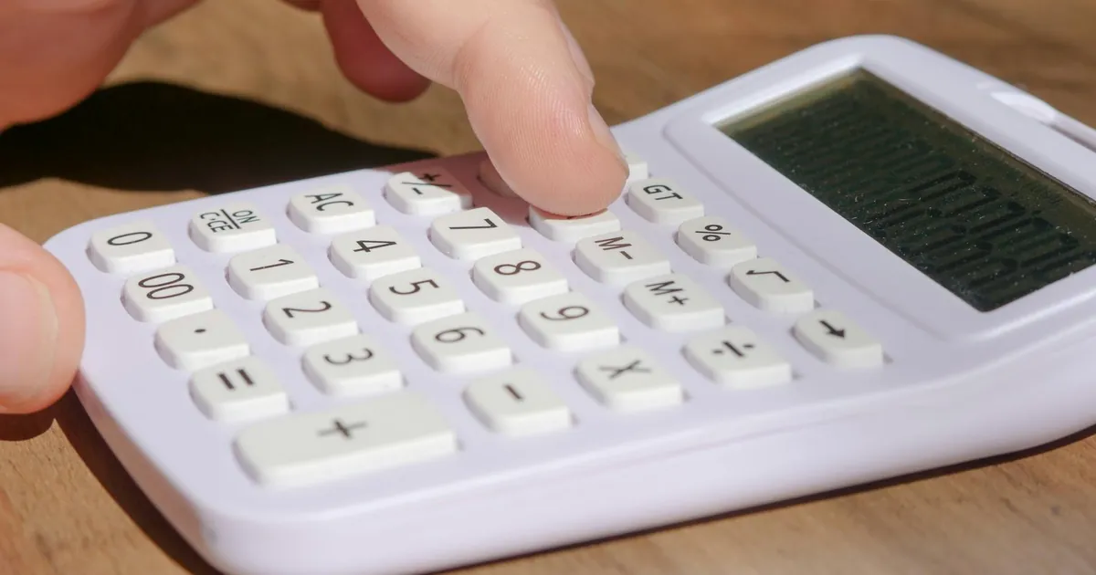 A hand pressing keys on a white calculator placed on a wooden desk in bright daylight.