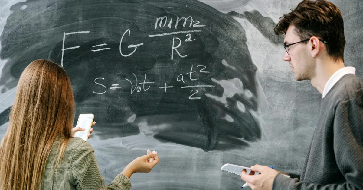 Two students discussing physics equations on a chalkboard in a classroom setting.