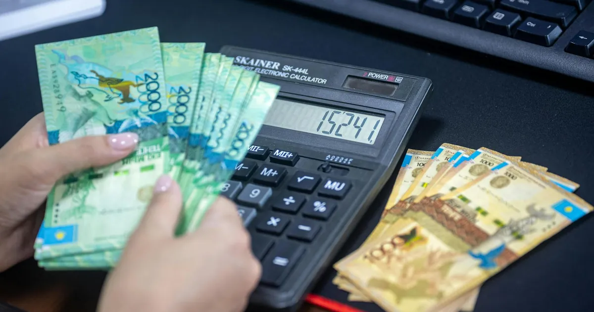Hands counting Kazakhstani tenge while using calculator at desk.