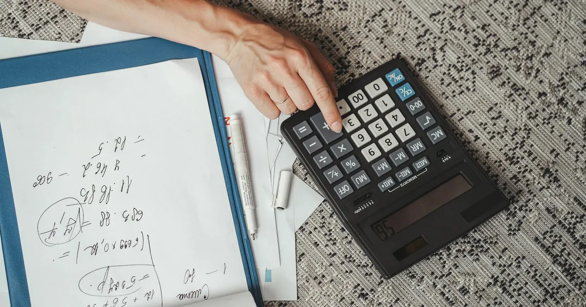Top view of a hand calculating numbers with a large calculator and notes on a desk.