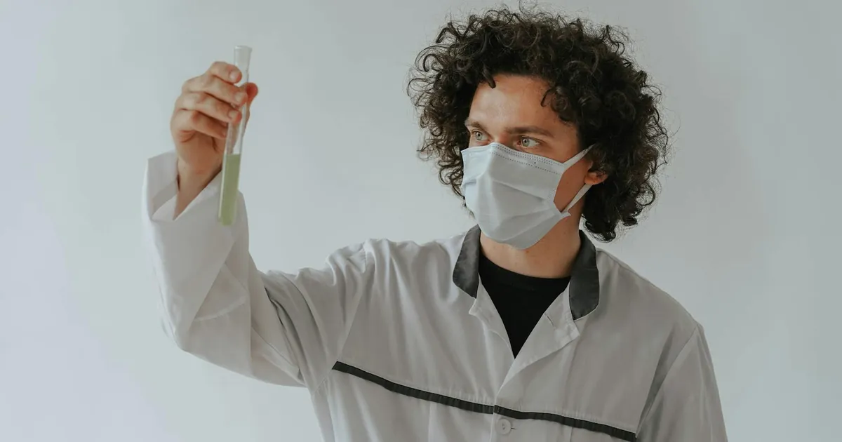 A scientist in a lab coat examining a test tube while wearing a face mask.