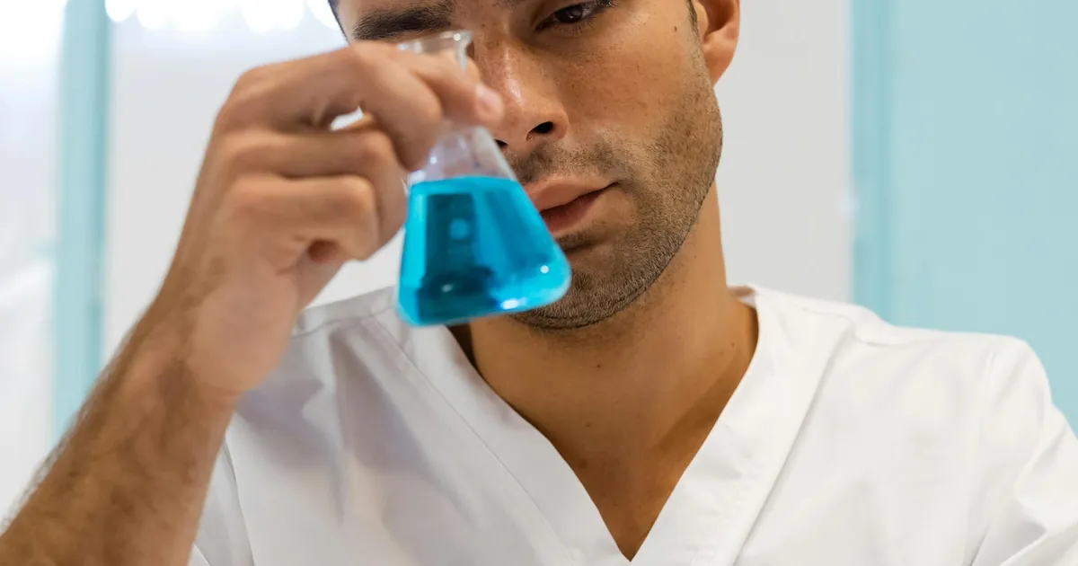 Scientist in lab coat examining blue liquid in flask, focused on experiment.