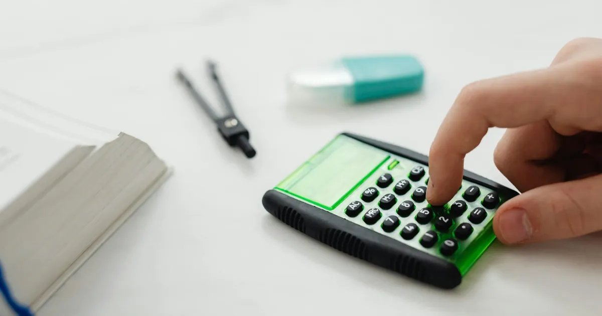 Close-up of a person's hand pressing a calculator on a desk with other stationery items.