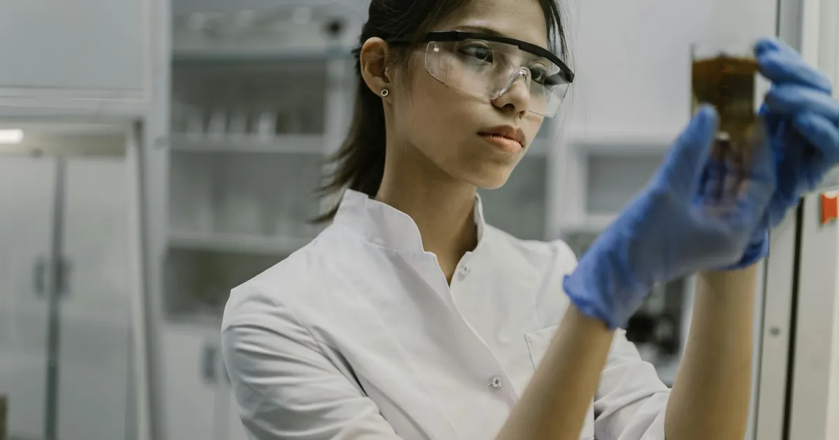 Young female scientist in lab coat examining a brown liquid in a glass beaker inside a laboratory.