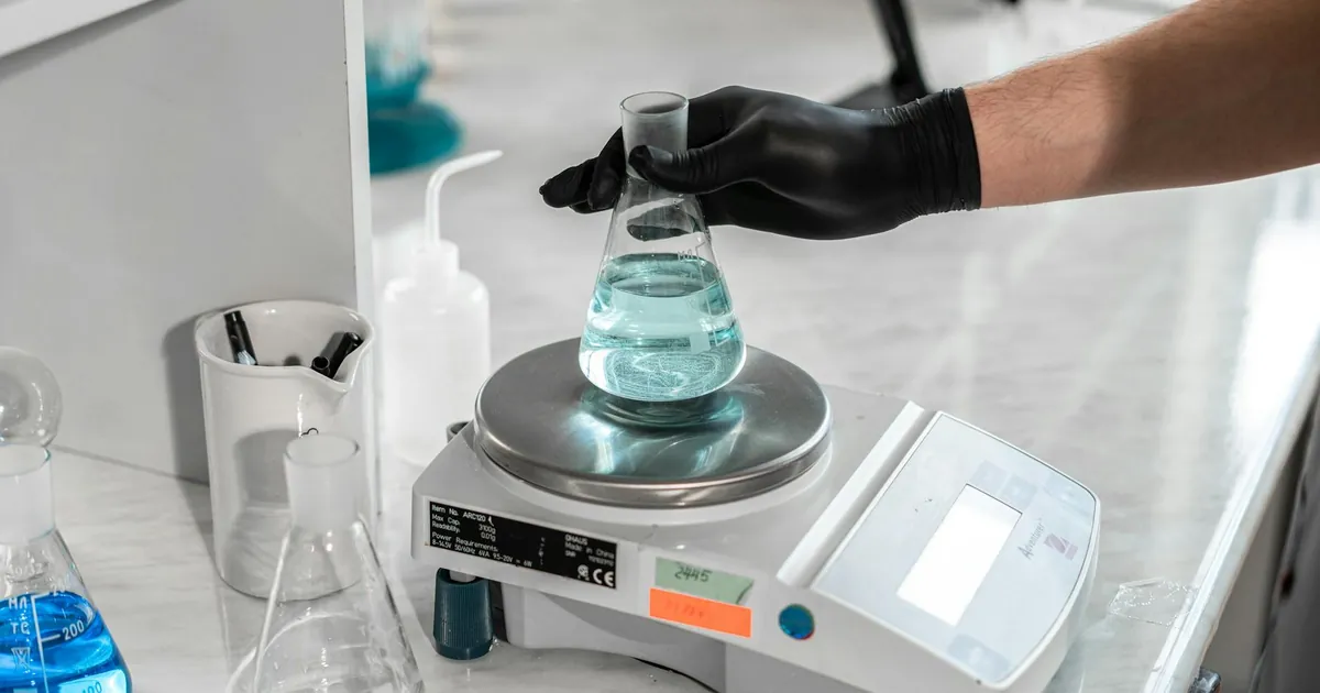 A scientist handling a liquid-filled flask on a laboratory scale for precise measurement.